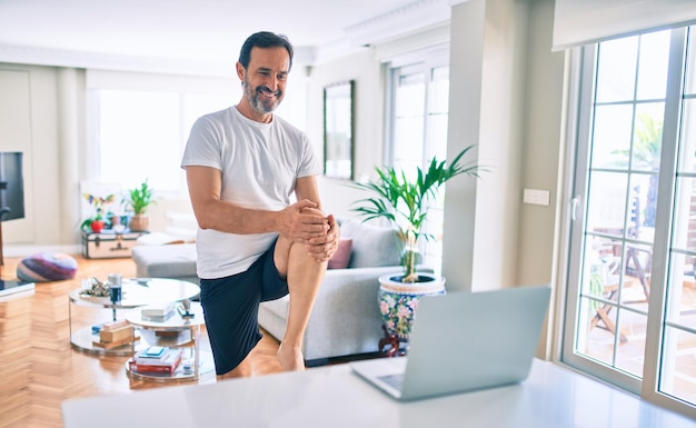 Homem de meia-idade com barba treinando e alongando fazendo exercícios em casa olhando para o vídeo do esporte no computador