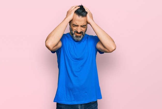 Homem de meia idade com barba vestindo camiseta casual azul sofrendo de dor de cabeça desesperado e estressado porque dor e enxaqueca mãos na cabeça