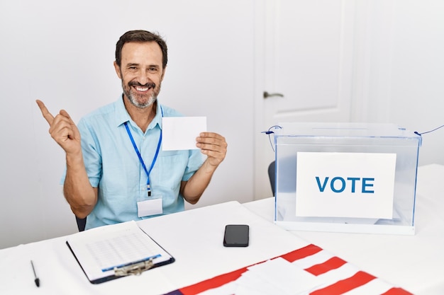 Homem de meia-idade com barba votando colocando envelope em urnas sorrindo feliz apontando com a mão e o dedo para o lado