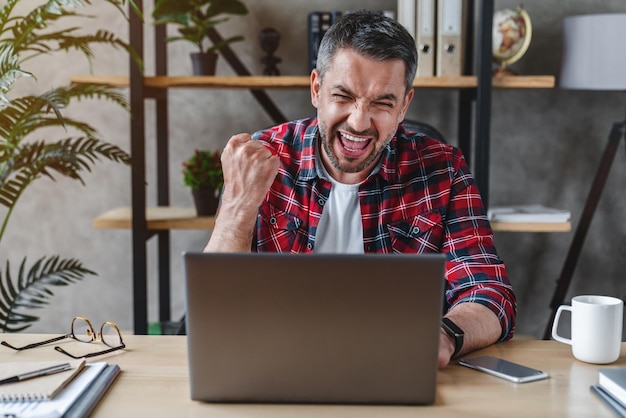 Homem de meia idade com braços levantados trabalhando no laptop e comemorando o sucesso em casa