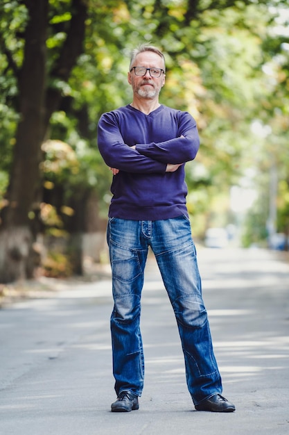 Homem de meia idade com cabelos grisalhos em um ambiente ao ar livre Suéter azul Óculos Barbudo empresário confiante Fundo desfocado Mãos cruzadas