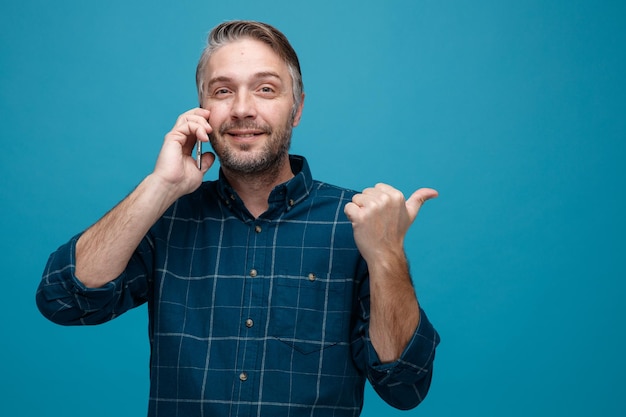 Homem de meia idade com cabelos grisalhos na camisa de cor escura falando no celular olhando para a câmera sorrindo apontando com o polegar para o lado em pé sobre fundo azul