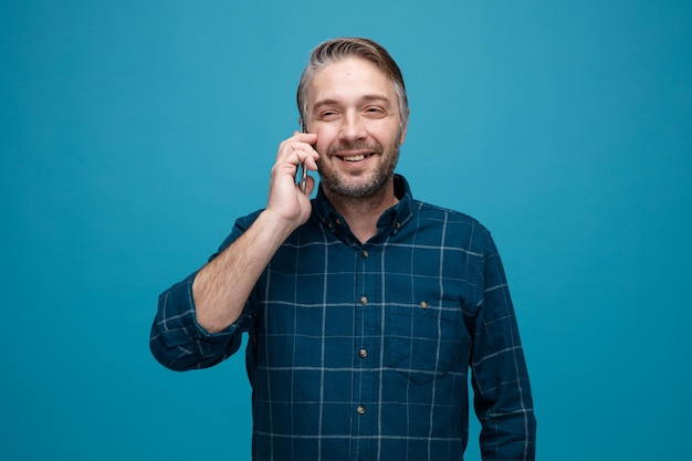 Homem de meia idade com cabelos grisalhos na camisa de cor escura falando no celular sorrindo alegremente feliz e positivo em pé sobre fundo azul