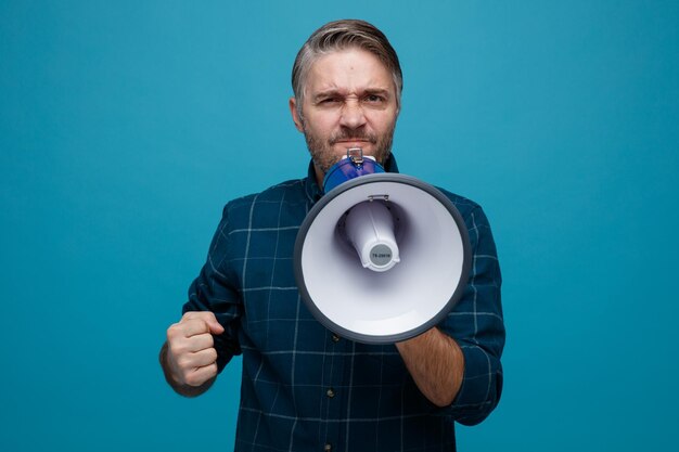 Foto homem de meia idade com cabelos grisalhos na camisa de cor escura, gritando no megafone com expressão agressiva, cerrando o punho em pé sobre fundo azul