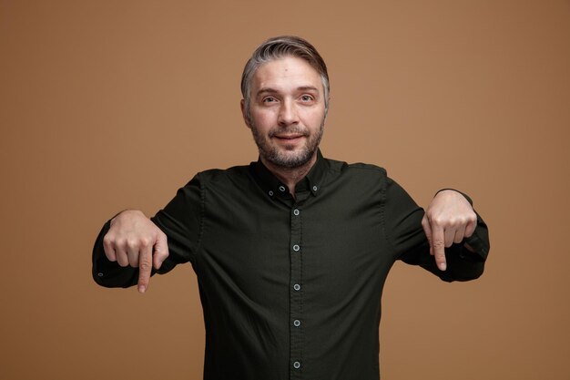 Foto homem de meia idade com cabelos grisalhos na camisa verde escura, olhando para a câmera sorrindo apontando com os dedos indicadores para baixo em pé sobre fundo marrom