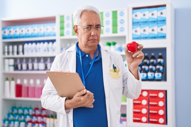 Homem de meia-idade com cabelos grisalhos, trabalhando na farmácia, segurando o coração vermelho, cético e nervoso, carrancudo, chateado por causa do problema da pessoa negativa