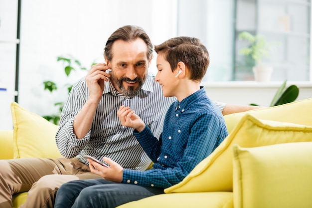 Homem de meia idade e barbudo alegre sentado com seu filho adolescente progressivo e sorrindo enquanto experimenta fones de ouvido sem fio