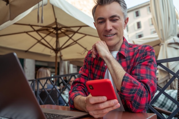 Homem de meia idade em camisa xadrez sentado à mesa no café da rua com um telefone nas mãos