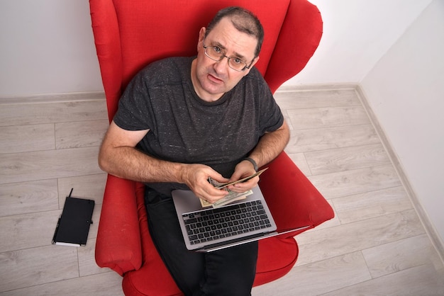 Homem de meia idade em óculos com um laptop no colo sentado em uma cadeira vermelha e contando dinheiro