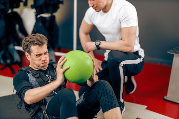 Homem de meia idade está fazendo exercícios abdominais durante o treinamento ems com personal trainer na academia.