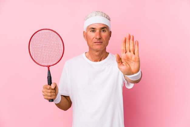 Homem de meia idade jogando badminton isolado em pé com a mão estendida, mostrando o sinal de pare, impedindo você.