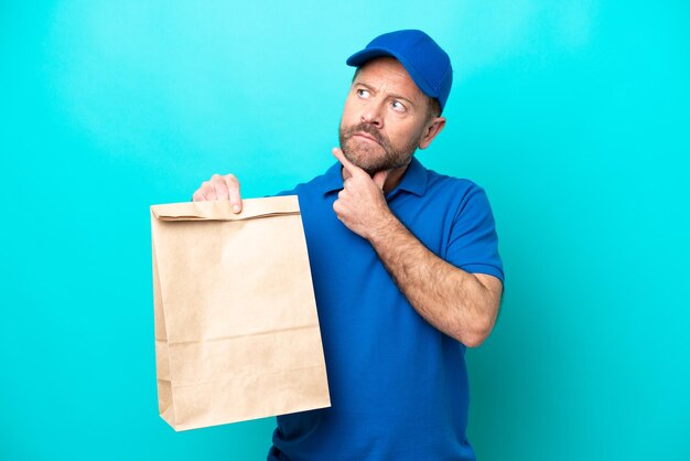 Homem de meia idade levando um saco de comida para viagem isolada em fundo azul com dúvidas