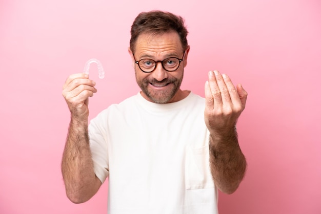 Homem de meia-idade segurando aparelho invisível isolado em fundo rosa convidando para vir com a mão feliz que você veio