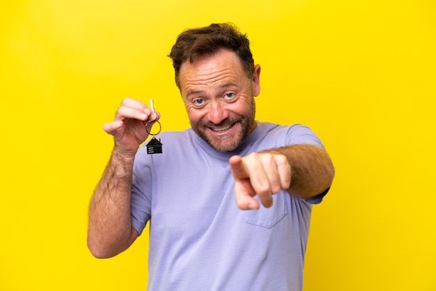 Foto homem de meia-idade segurando as chaves de casa isoladas em fundo amarelo aponta o dedo para você com uma expressão confiante