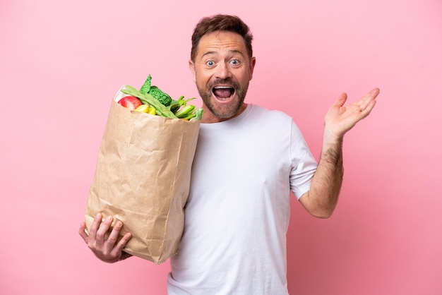 Homem de meia-idade segurando uma sacola de compras isolada em fundo rosa com expressão facial chocada