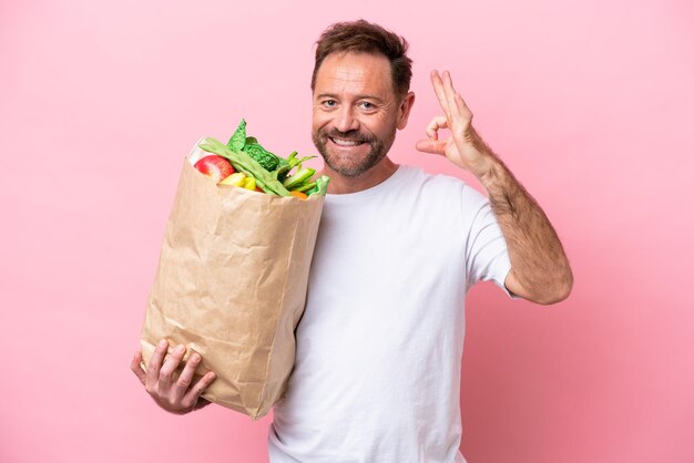 Homem de meia-idade segurando uma sacola de compras isolada em fundo rosa mostrando sinal de ok com os dedos