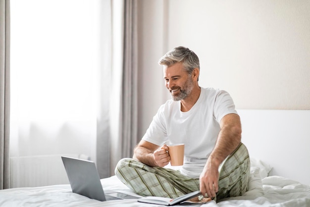 Homem de meia idade sentado na cama com laptop e bloco de notas