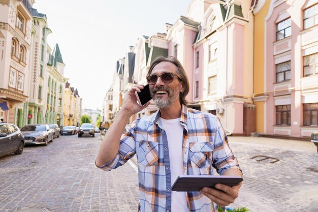 Homem de meia idade sorridente e feliz, com barba grisalha, falando ao telefone, segurando o tablet digital na mão, ao ar livre, no fundo da cidade velha, vestindo camisa xadrez e óculos de sol Freelancer, homem viajante