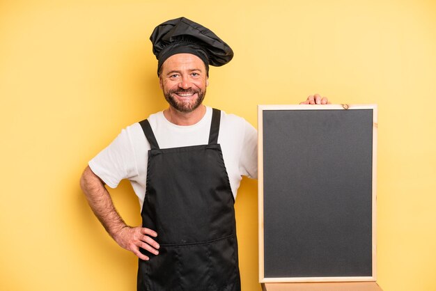Homem de meia idade sorrindo alegremente com uma mão no quadril e confiante. conceito de chef e quadro-negro