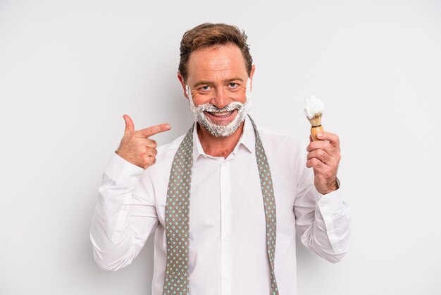 homem de meia idade sorrindo com confiança apontando para o próprio sorriso largo. pincel de barbear e conceito de espuma