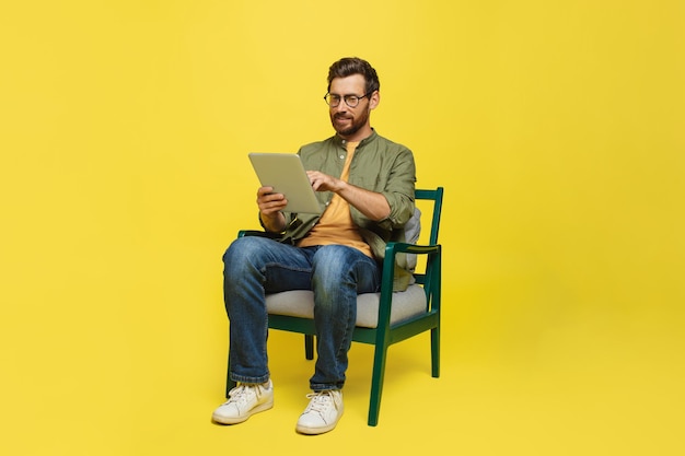 Homem de meia idade trabalhando em tablet digital sentado na cadeira sobre o espaço de cópia de fundo de estúdio amarelo