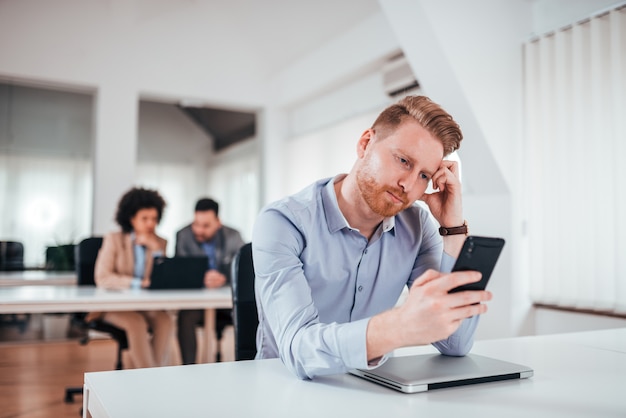 Homem de negócio cansado que olha o telefone em seu local de trabalho.