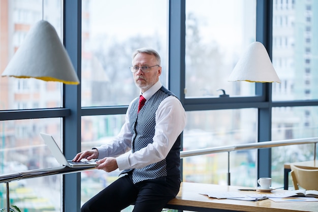 Homem de negócios adulto, professor, mentor trabalhando em um novo projeto. Senta-se junto a uma grande janela sobre a mesa. Ele olha para a tela do laptop.