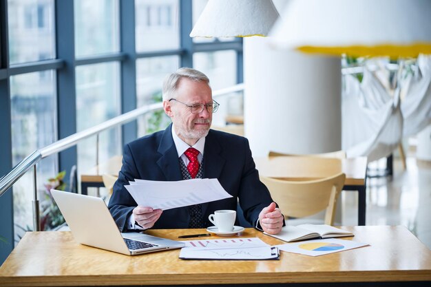 Homem de negócios adulto trabalhando em um novo projeto e olhando os gráficos de crescimento das ações. Senta-se em uma grande janela na mesa. Olha para a tela do laptop e bebe café.