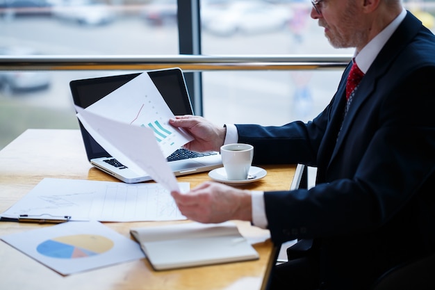 Homem de negócios adulto trabalhando em um novo projeto e olhando os gráficos de crescimento das ações. senta-se em uma grande janela na mesa. olha para a tela do laptop e bebe café.