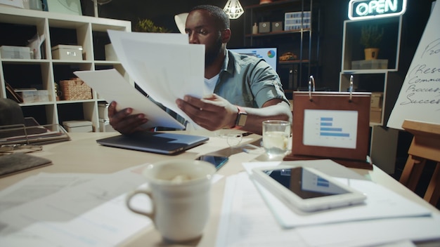 Homem de negócios africano pensativo segurando gráficos financeiros no coworking hipster