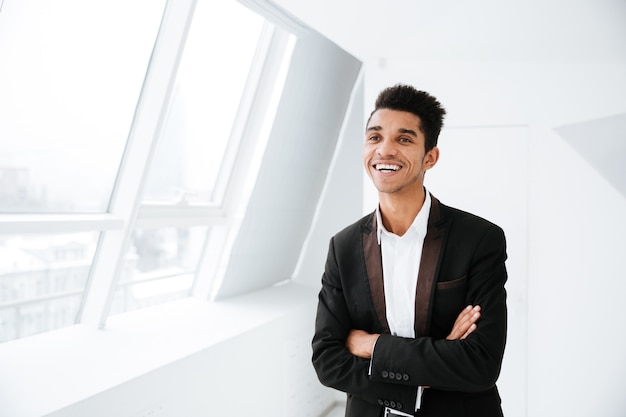 Homem de negócios africano sorridente de terno preto em pé perto da janela no escritório com os braços cruzados e olhando para o lado
