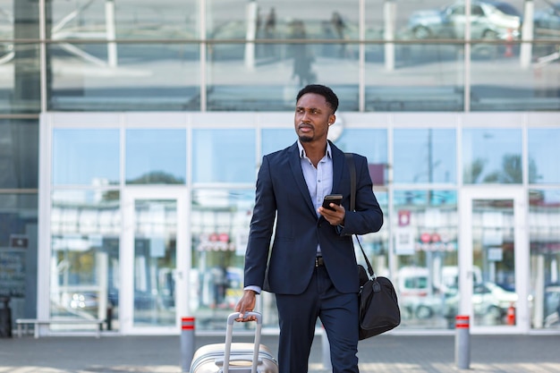 Homem de negócios africano viajando perto do aeroporto com uma mala chamando um táxi usando um aplicativo móvel