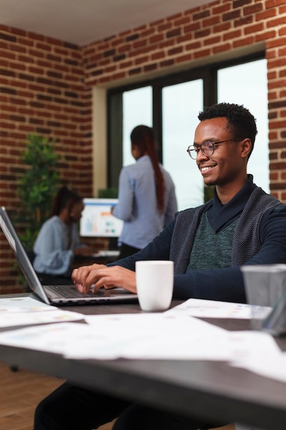 Homem de negócios afro-americano sorridente desenvolvendo estratégia de marketing de inicialização e plano de propaganda. funcionário do escritório da empresa trabalhando em dados gráficos financeiros enquanto usa o laptop do computador de trabalho.