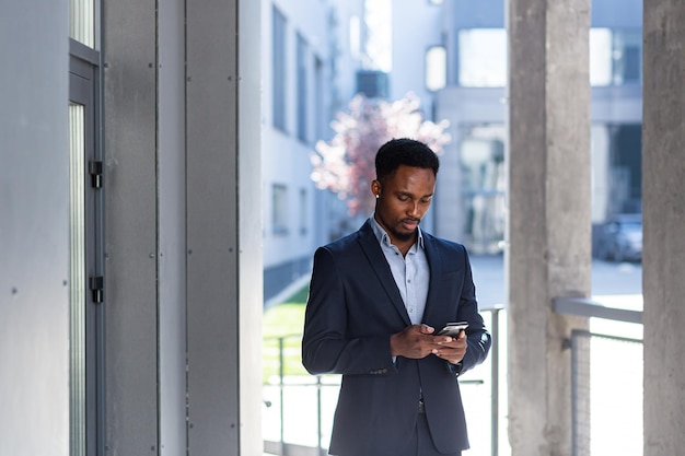 Homem de negócios afro-americano trabalhando em videochamada usa telefone e aplicativo, fora do escritório, homem alegre e bem-sucedido em um terno de negócios