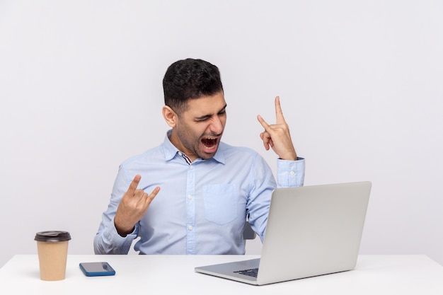 Homem de negócios alegre despreocupado sentado no local de trabalho do escritório, mostrando o gesto com a mão do rock and roll, sinal de chifre do diabo, gritando louco enquanto fala em videochamada no laptop. tiro de estúdio isolado no fundo branco