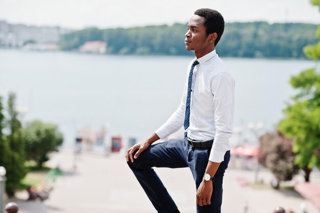 Homem de negócios americano africano elegante em calças e camisa posou ao ar livre contra o lago