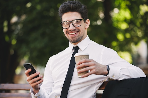 Homem de negócios ao ar livre no parque usando telefone celular, bebendo café.