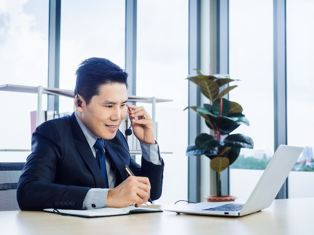 Homem de negócios asiático de terno com fone de ouvido usando laptop na mesa no escritório. Homem com fones de ouvido e microfone fazendo anotações e usando laptop com teleconferência