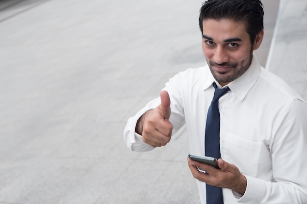 Homem de negócios asiático feliz usando telefone inteligente com o polegar para cima; retrato de um homem de negócios asiático do norte da Índia com um sorriso feliz e sorridente usando um telefone inteligente para app, conexão de internet, conceito de boa conexão