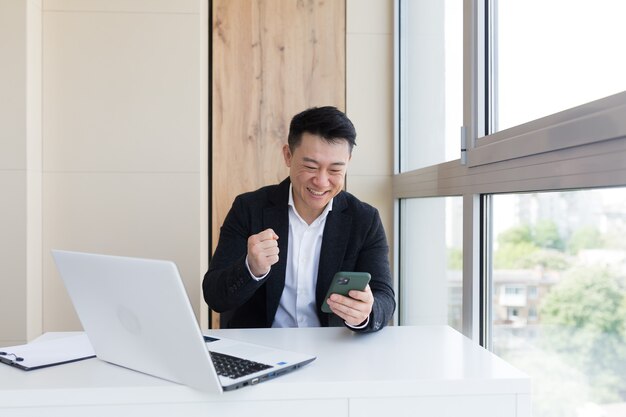 Homem de negócios asiático no escritório olhando para o celular com o vencedor ou vitória de emoção