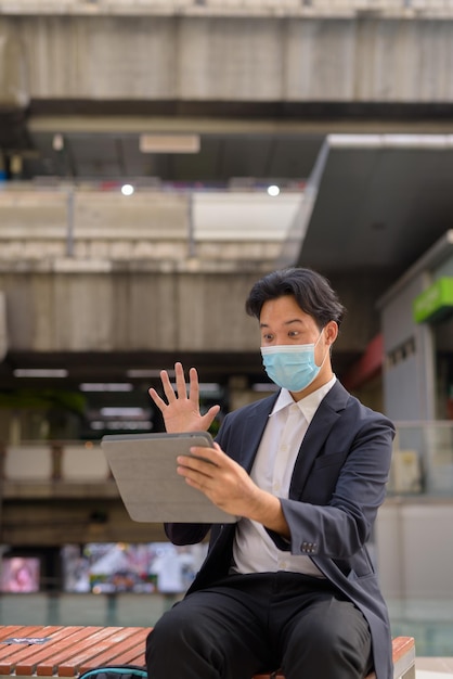 Homem de negócios asiático usando máscara facial e sentado ao ar livre na cidade usando um computador tablet digital