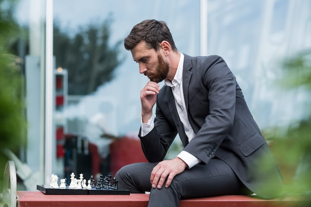 Homem de negócios banqueiro jogando xadrez, sentado no banco perto do escritório, pensativo, fazendo um movimento estratégico