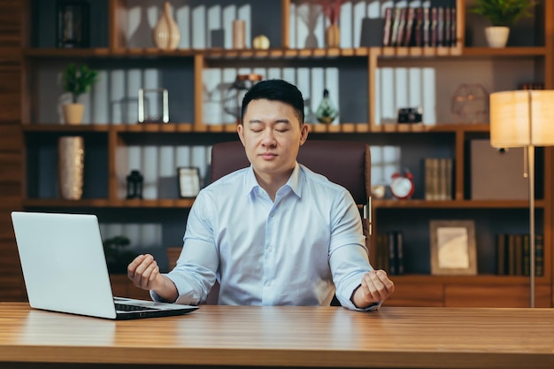 Homem de negócios bem sucedido medita sentado em uma mesa em um escritório clássico homem asiático de camisa relaxa depois de trabalhar no laptop no trabalho
