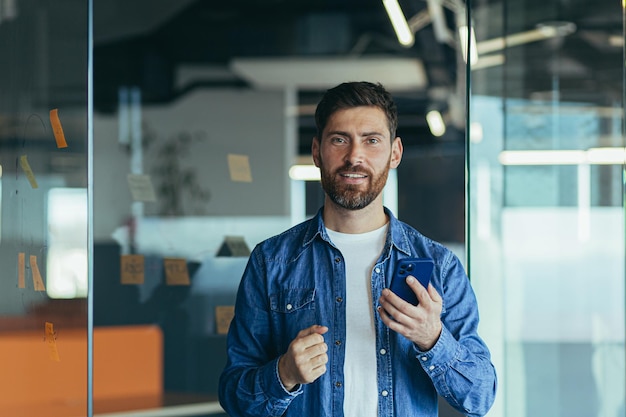 Homem de negócios bem-sucedido no escritório usando smartphone e sorrindo Empresário feliz usando aplicativos móveis enviando mensagens de texto navegando na internet olhando para smartphone sênior trabalhando com dispositivos móveis