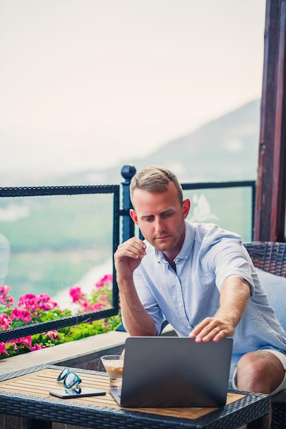 Homem de negócios bem sucedido trabalhando de férias atrás de um laptop com vista para a montanha Fluxo de trabalho do gerente online Trabalhe ao ar livre com uma bela vista da varanda