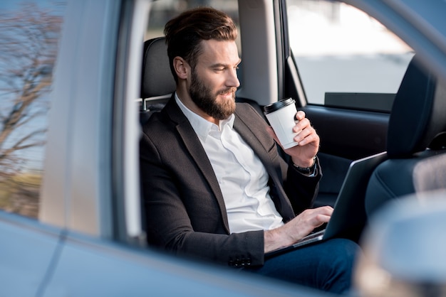 Homem de negócios bonitão sentado tomando café no banco de trás do carro