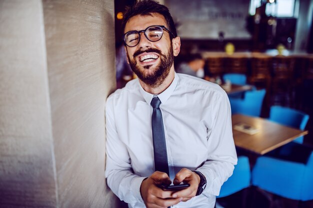 Homem de negócios bonito barbudo caucasiano elegante na camisa, gravata e com óculos encostado na parede no café e usando o telefone inteligente para ler ou enviar mensagem.