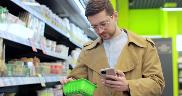 Homem de negócios bonito com carrinho de compras usando a lista de verificação de comida do smartphone andando dentro da mercearia