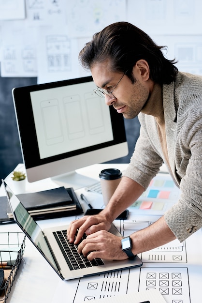 Homem de negócios bonito concentrado de óculos em pé na mesa com esquemas de design de aplicativos e digitando no laptop no escritório