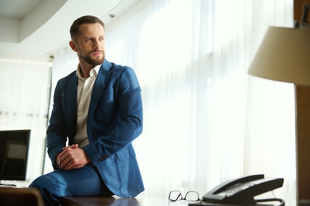 Foto homem de negócios bonito confiante, homem atraente, investidor financeiro próspero em viagem de negócios, sentado à mesa do escritório em um quarto de hotel e olhando pensativamente de lado no fundo das janelas do quarto de hotel